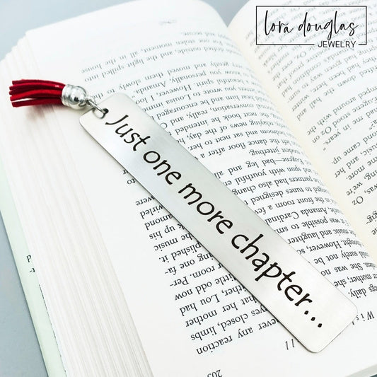 A stainless steel metal bookmark with the engraved text 'Just One More Chapter' and a tassel, placed on an open book.