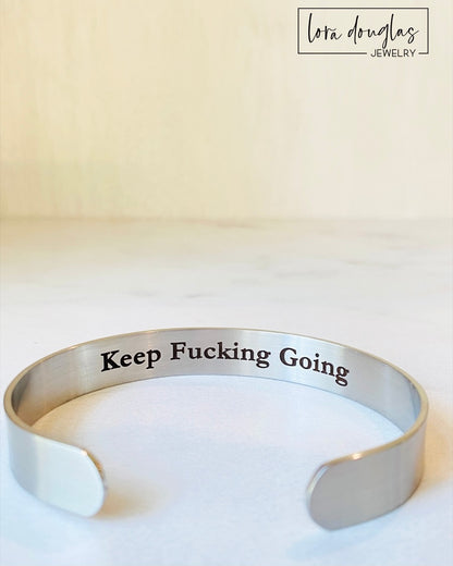A stainless steel cuff bracelet with the phrase 'KEEP FUCKING GOING' engraved on the inside, against the skin side, in a clear, bold typeface.