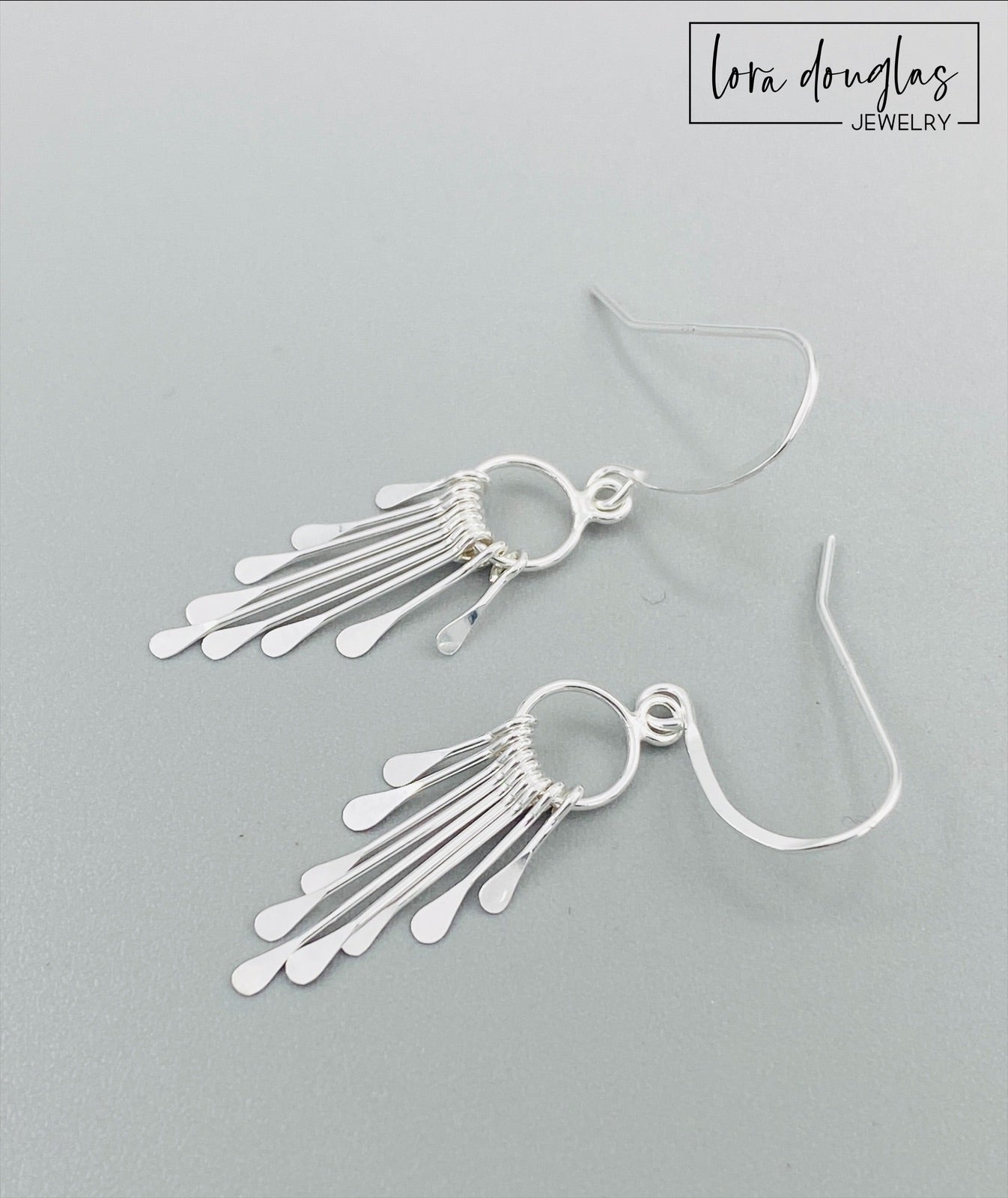A pair of sterling silver fringe paddle earrings with a drop-style design, featuring small fringes dangling from a main hoop.