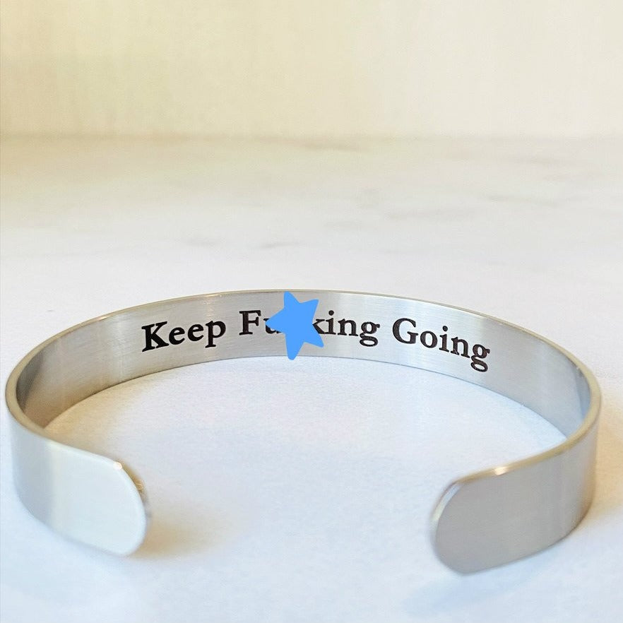 A stainless steel cuff bracelet with the phrase 'KEEP FUCKING GOING' engraved on the inside, against the skin side, in a clear, bold typeface.