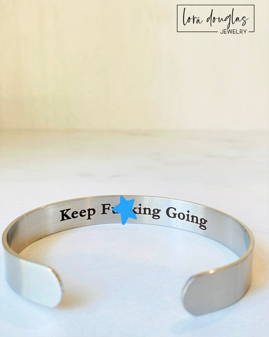 A stainless steel cuff bracelet with the phrase 'KEEP FUCKING GOING' engraved on the inside, against the skin side, in a clear, bold typeface.