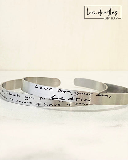 A stainless steel cuff bracelet with engraved handwriting on it, displayed on a light background.