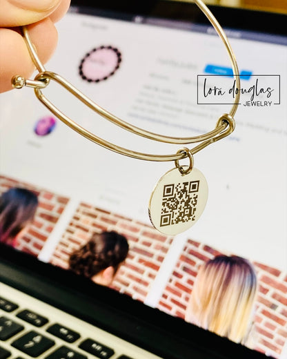 A QR code charm placed on a display stand, with a silver chain necklace that says 'Lost - will you tell my family I'm ok?' engraved on a blue disc.