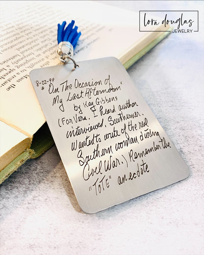 A custom engraved bookmark with a handwritten message engraved on the metal. The bookmark is silver with black engraving and has a navy blue Tassel.