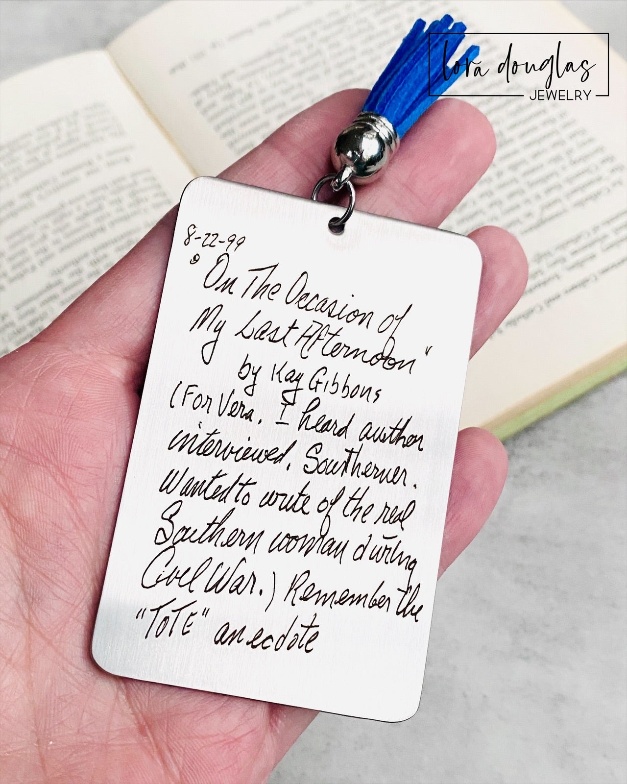 A custom engraved bookmark with a handwritten message engraved on the metal. The bookmark is silver with black engraving and has a navy blue Tassel.