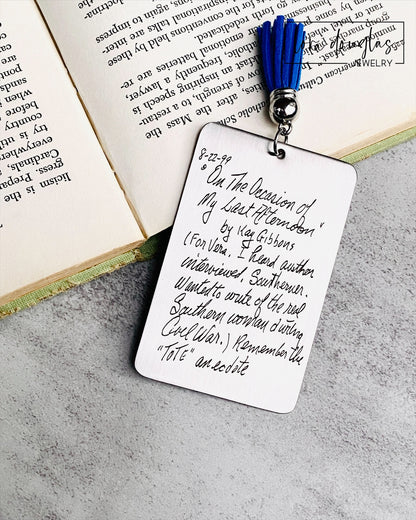A custom engraved bookmark with a handwritten message engraved on the metal. The bookmark is silver with black engraving and has a navy blue Tassel.