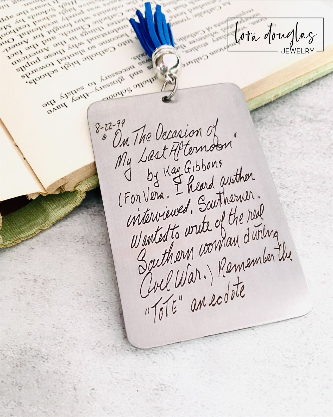 A custom engraved bookmark with a handwritten message engraved on the metal. The bookmark is silver with black engraving and has a navy blue Tassel.