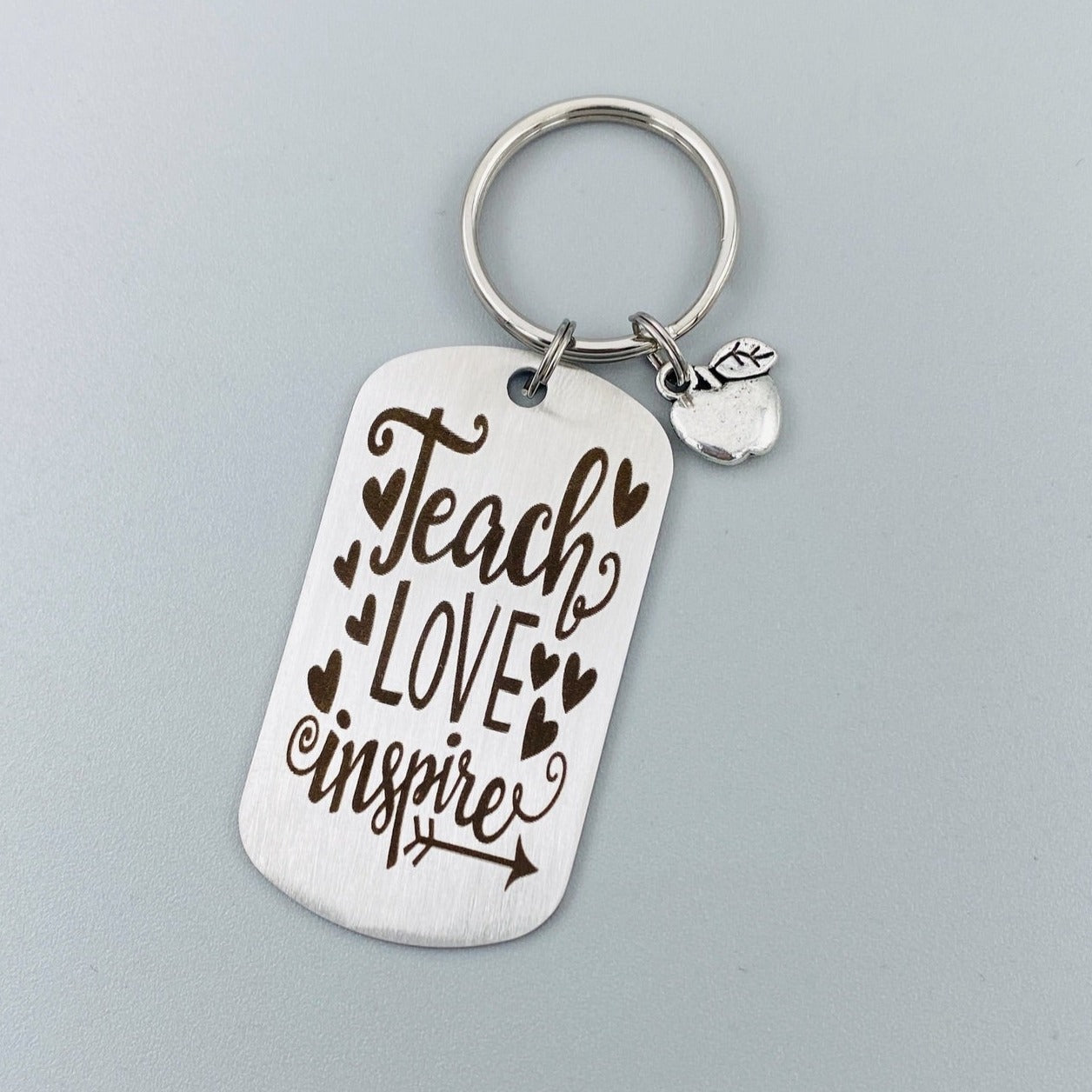 A silver stainless steel dog tag keychain with a silver pewter apple charm, engraved with the text 'Teach Love Inspire' accompanied by a heart symbol.
