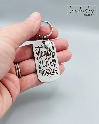 A silver stainless steel dog tag keychain with a silver pewter apple charm, engraved with the text 'Teach Love Inspire' accompanied by a heart symbol.