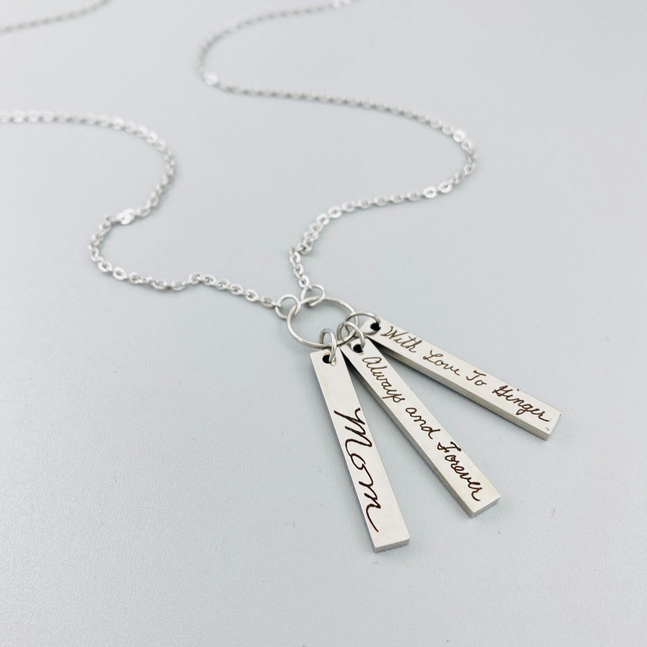 A silver stainless steel necklace with a chain and three rectangular pendants, each engraved with different handwriting.