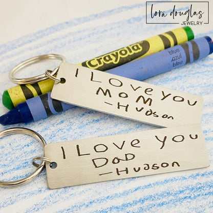 A stainless steel keychain with engraved handwriting samples. The keychain features a silver-colored plate with the text 'I love you dad' and 'Mom you're the best' alongside a standard key ring.