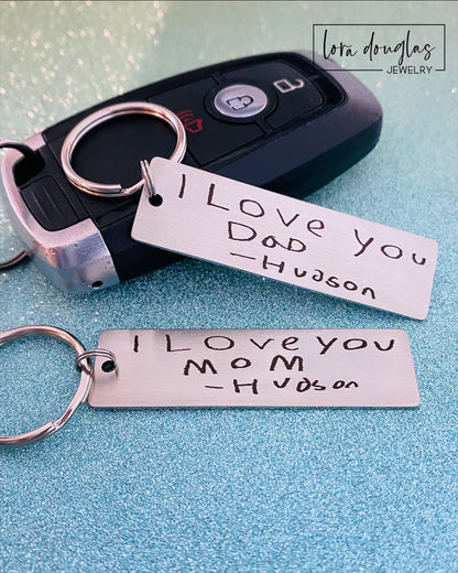 A stainless steel keychain with engraved handwriting samples. The keychain features a silver-colored plate with the text 'I love you dad' and 'Mom you're the best' alongside a standard key ring.