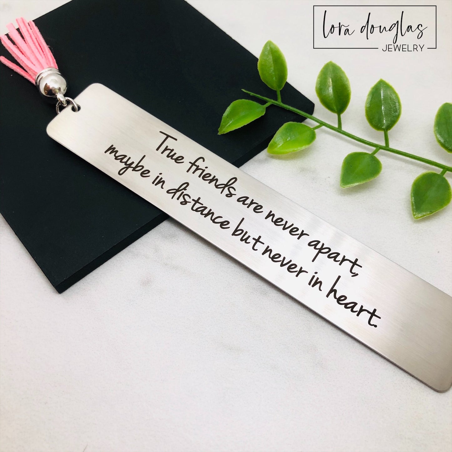 engraved metal bookmark with friendship quote displayed inside open novel pages, personalized long distance friendship gift featuring brushed stainless steel finish and colorful tassel options