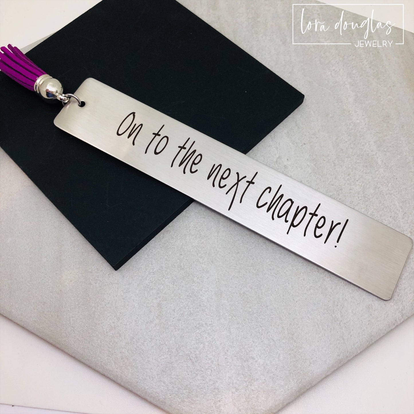 engraved on to the next chapter bookmark displayed beside stacked books and tassel, stainless steel inspirational bookmark gift for graduates teachers retirees and book lovers