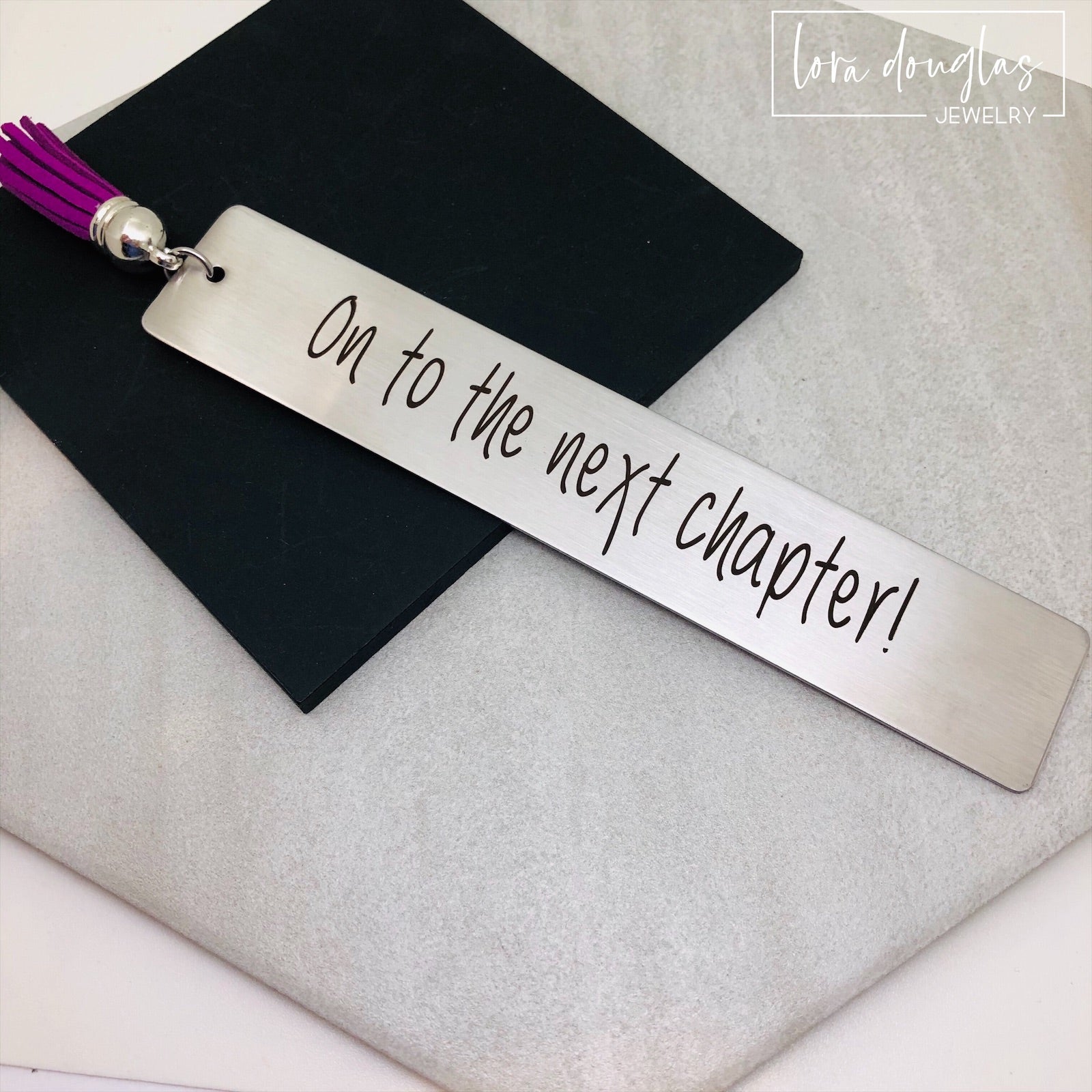 engraved on to the next chapter bookmark displayed beside stacked books and tassel, stainless steel inspirational bookmark gift for graduates teachers retirees and book lovers