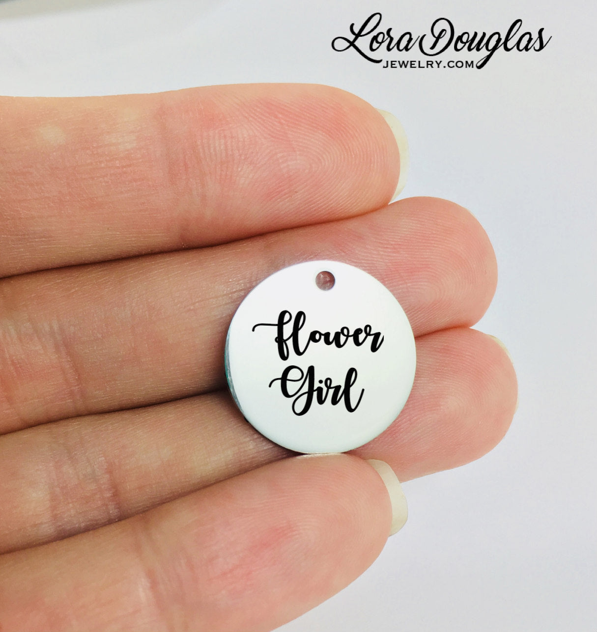 A round stainless steel pendant with the engraved text 'Flower Girl' held between fingers.