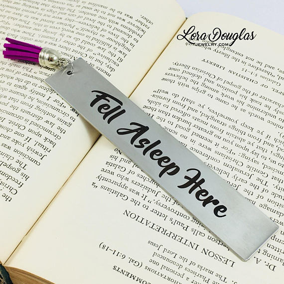 A stainless steel bookmark with the text 'Fell Asleep Here' engraved on it, displayed on an open book. The bookmark has a tassel and is attached to a navy blue velvet drawstring pouch with a gift tag.