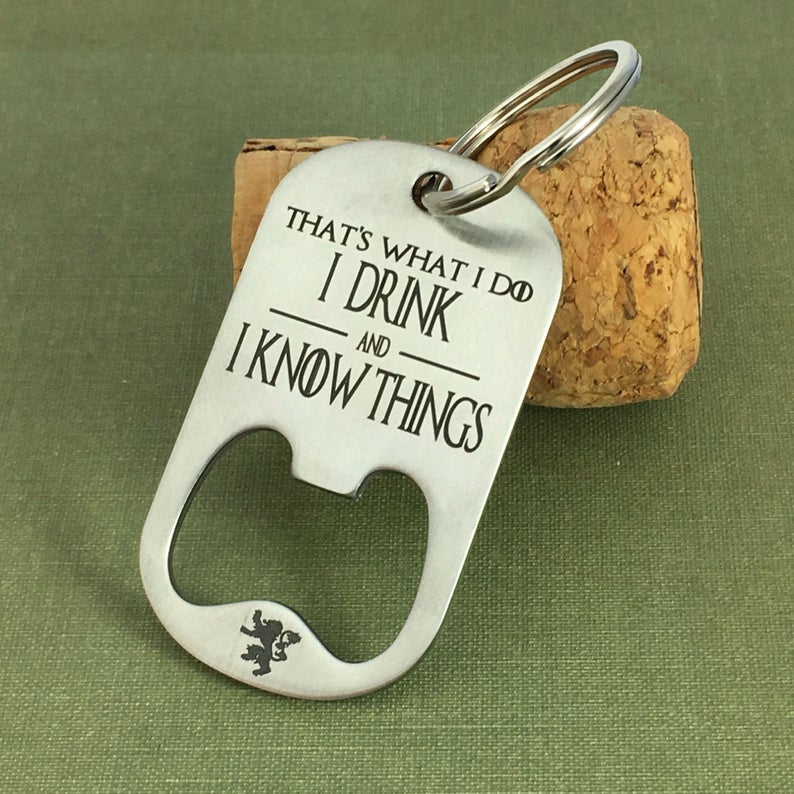 A stainless steel key chain with the engraved text 'That's What I Do I Drink and I Know Things' along with the HBO logo for Game of Thrones, resting on a corkscrew.