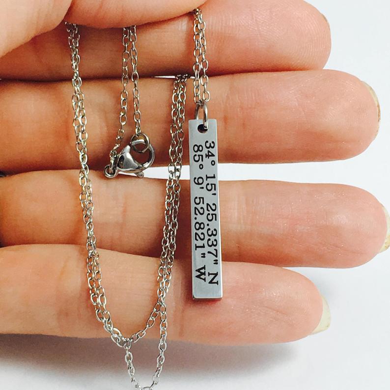 A custom coordinates necklace with a silver stainless steel pendant and chain, displayed against a floral background.