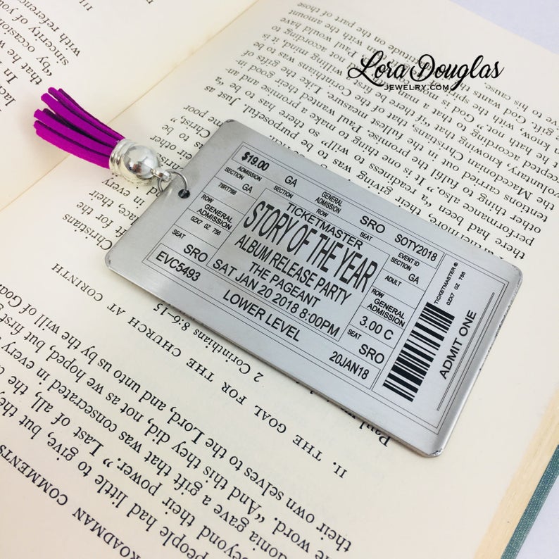 A stainless steel bookmark with laser engraved personalized text, depicting a concert ticket design, placed on a surface with a black bookmark and a green leaf in the background.