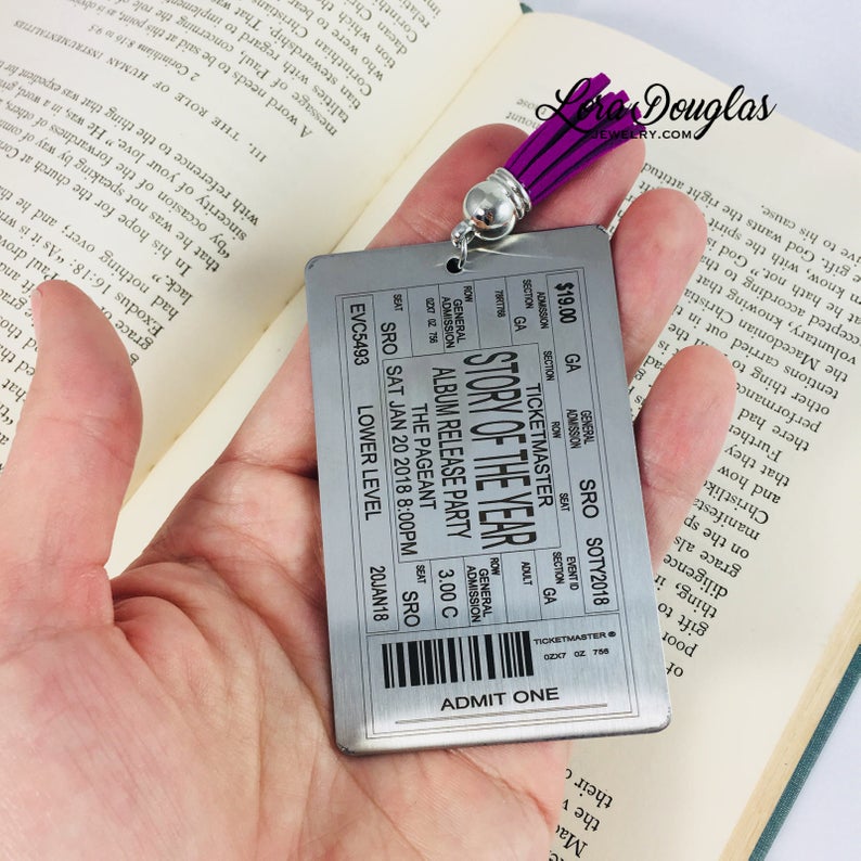 A stainless steel bookmark with laser engraved personalized text, depicting a concert ticket design, placed on a surface with a black bookmark and a green leaf in the background.