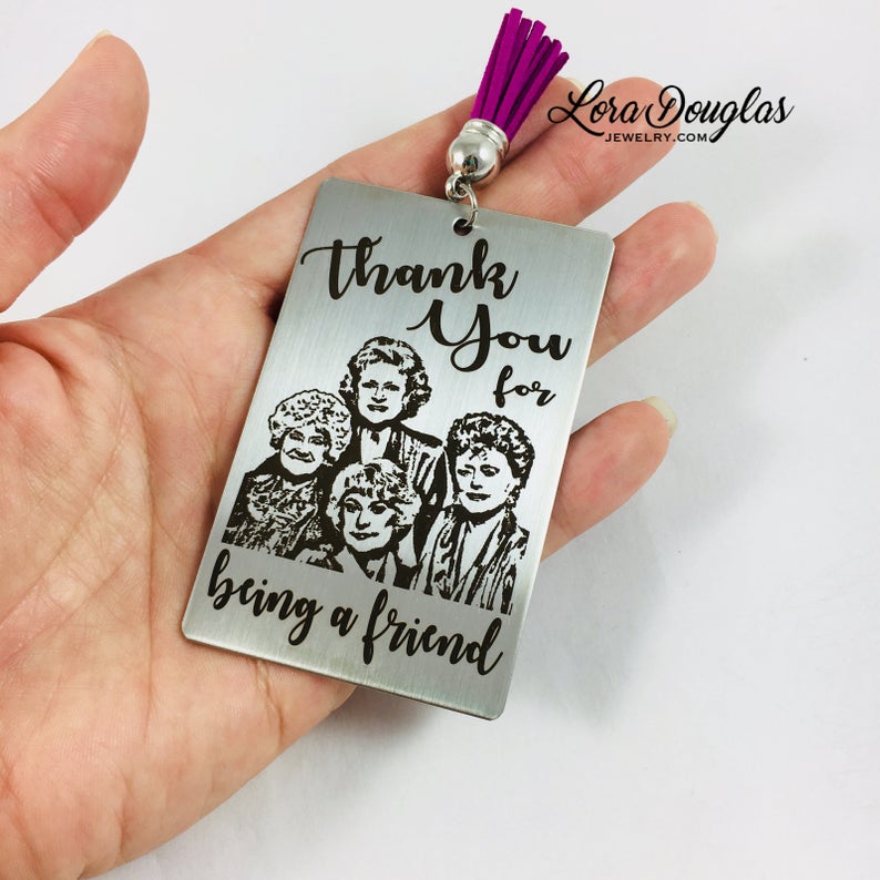 A stainless steel bookmark with engraved text and illustrations of the Golden Girls characters, with a navy blue velvet drawstring pouch and a gift tag.