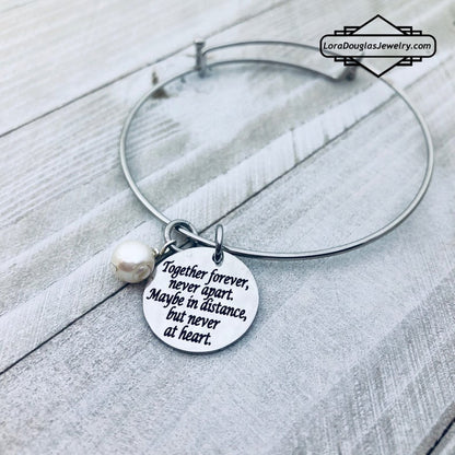 A stainless steel adjustable bangle bracelet with a round pendant that has engraved text 'Together Forever, Never Apart, Maybe in Distance, But Never at Heart'.
