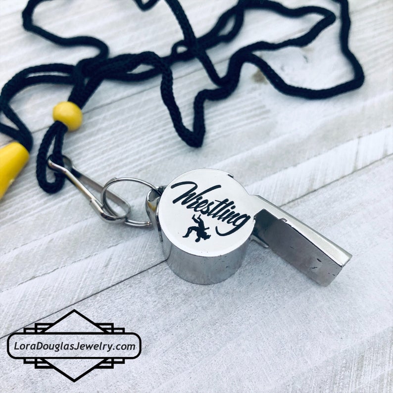 personalized stainless steel whistles with engraved text, one with 'COACH CARLOS' and sports icons, the other with 'FOOTBALL BASKETBALL SOCCER LIFEGUARD AND MORE'. Both whistles have a lanyard 