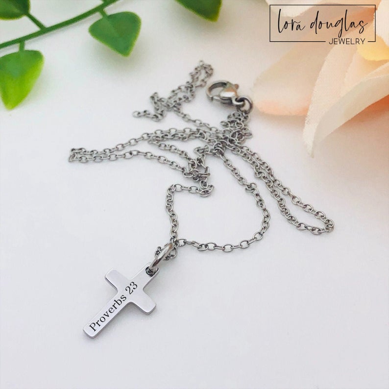 A silver stainless steel engraved cross necklace with a lobster clasp and a small cross charm, placed on a white surface with a few green leaves and a flower for decoration.