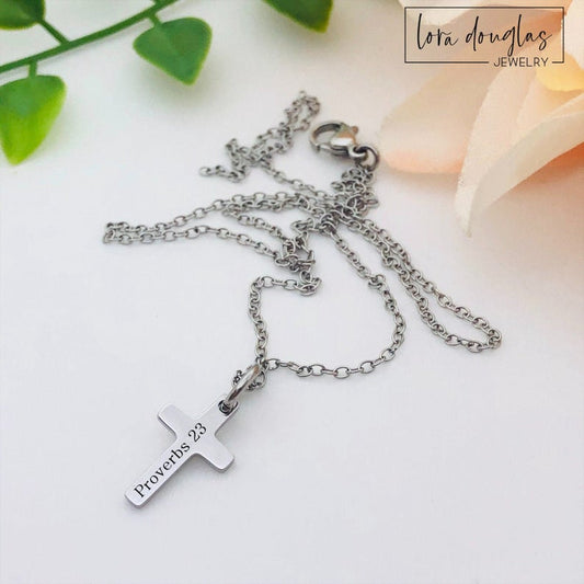 A silver stainless steel engraved cross necklace with a lobster clasp and a small cross charm, placed on a white surface with a few green leaves and a flower for decoration.