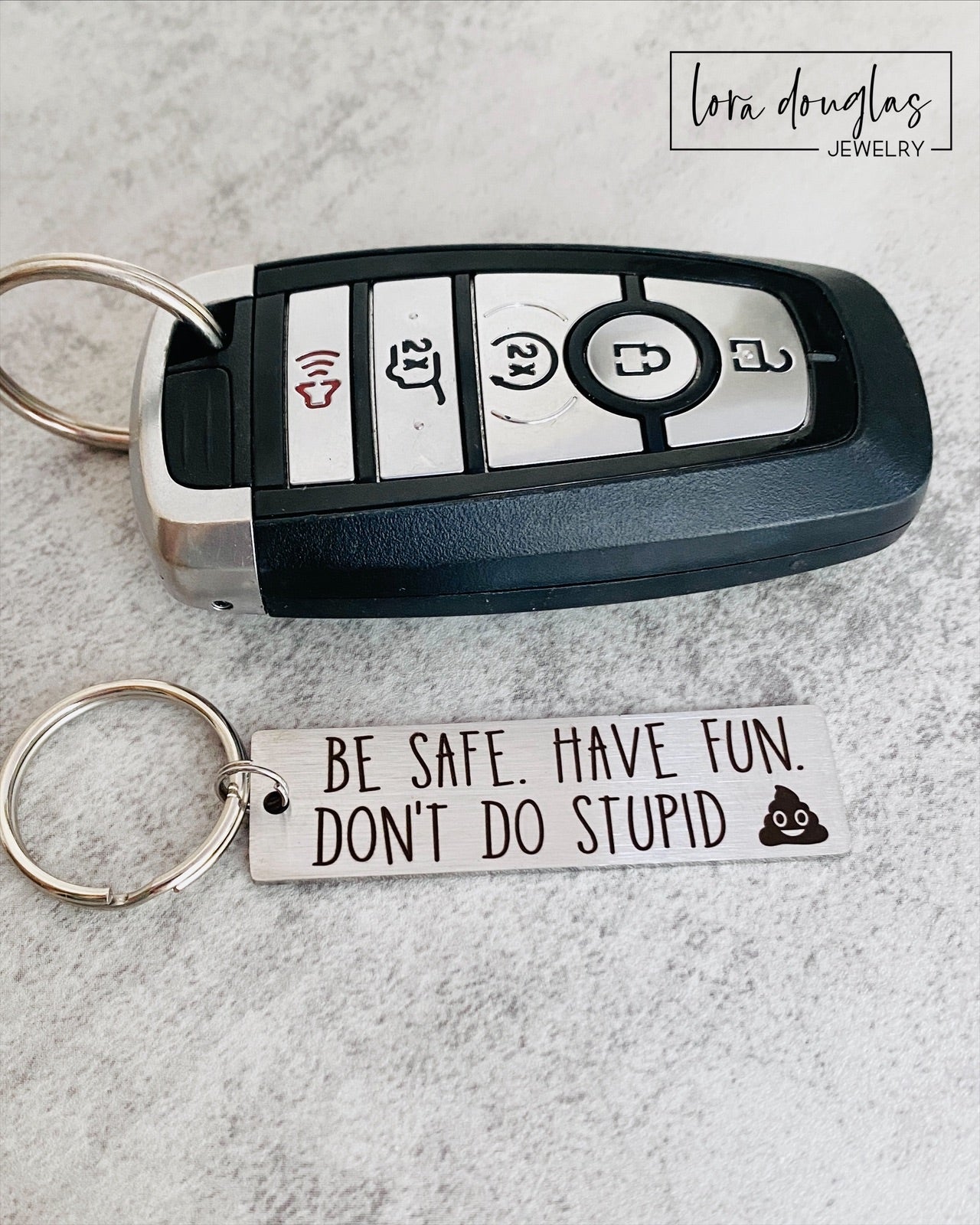 A metal keychain with an engraved message BE SAFE HAVE FUN DONT DO STUPID accompanied by a poop emoji, placed on a car key.