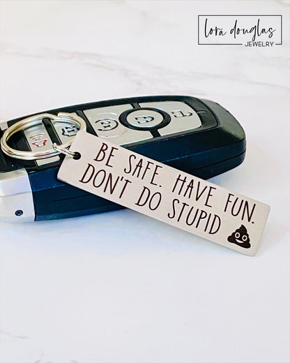 A metal keychain with an engraved message BE SAFE HAVE FUN DONT DO STUPID accompanied by a poop emoji, placed on a car key.