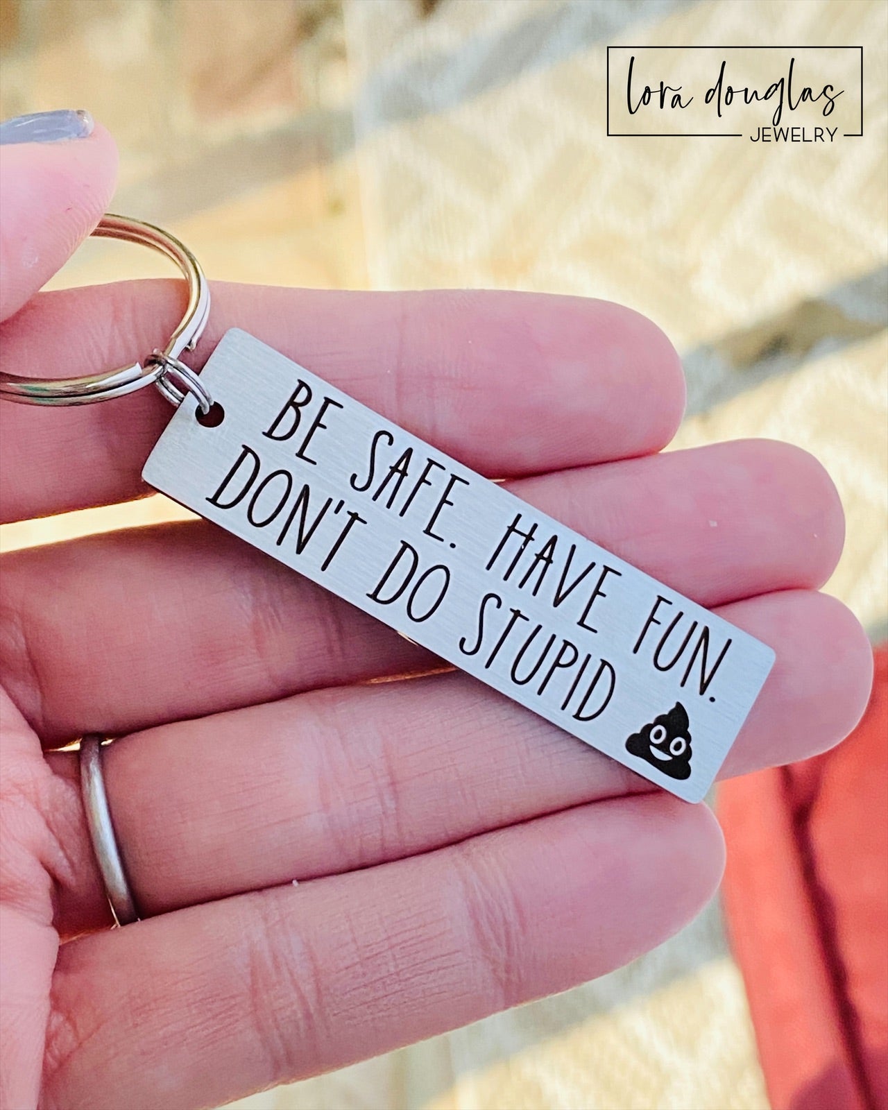 A metal keychain with an engraved message BE SAFE HAVE FUN DONT DO STUPID accompanied by a poop emoji, placed on a car key.