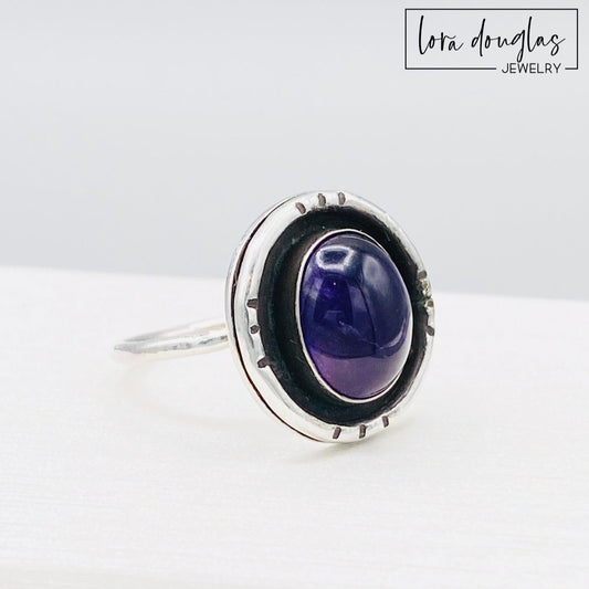 A sterling silver shadow box ring with a central amethyst stone, set in a circular silver setting.