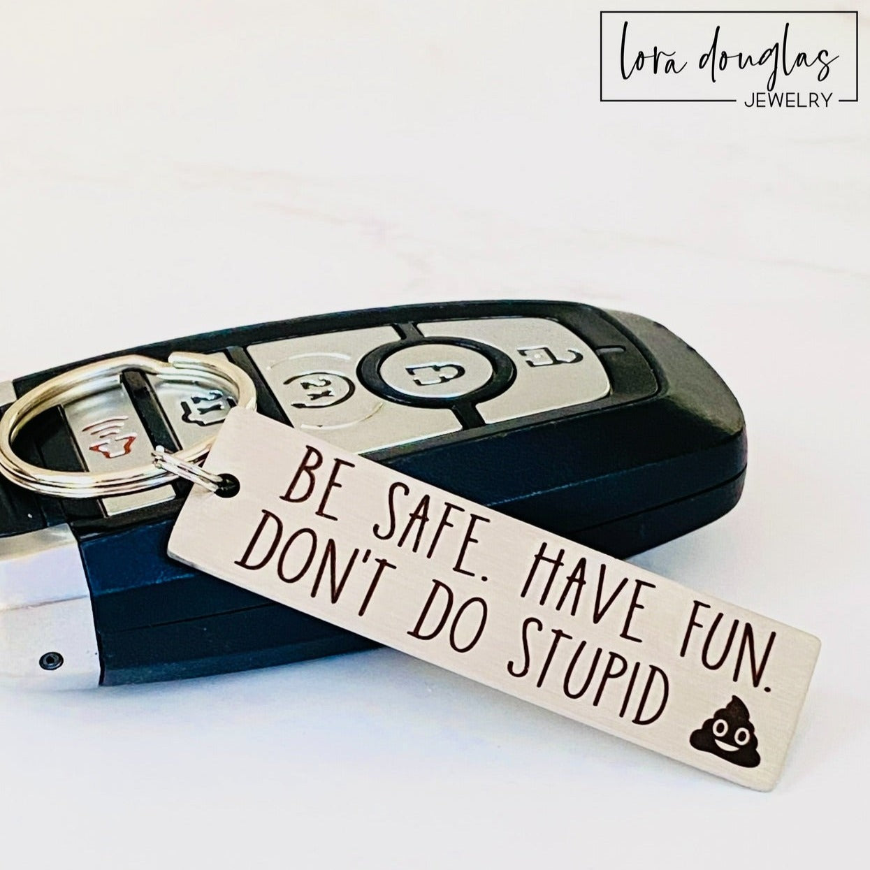 A metal keychain with an engraved message BE SAFE HAVE FUN DONT DO STUPID accompanied by a poop emoji, placed on a car key.