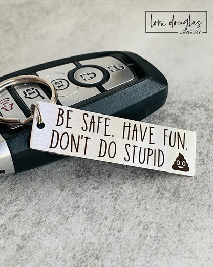 A metal keychain with an engraved message BE SAFE HAVE FUN DONT DO STUPID accompanied by a poop emoji, placed on a car key.