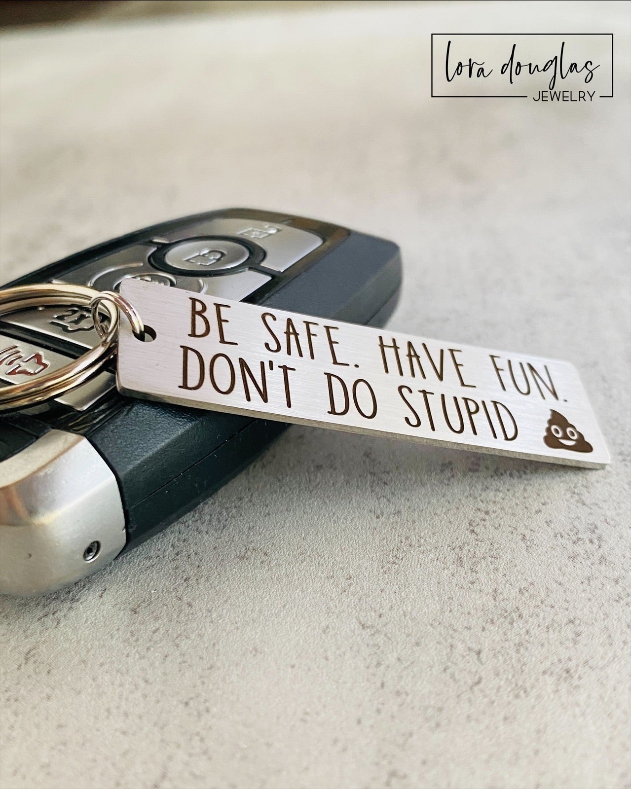 A metal keychain with an engraved message BE SAFE HAVE FUN DONT DO STUPID accompanied by a poop emoji, placed on a car key.