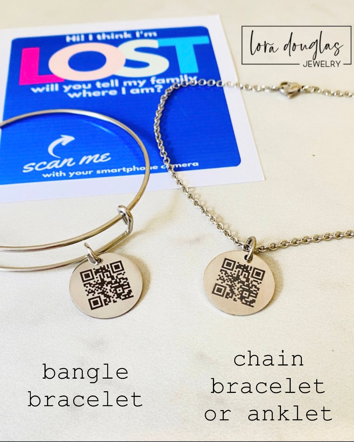 A QR code charm placed on a display stand, with a silver chain necklace that says 'Lost - will you tell my family I'm ok?' engraved on a blue disc.