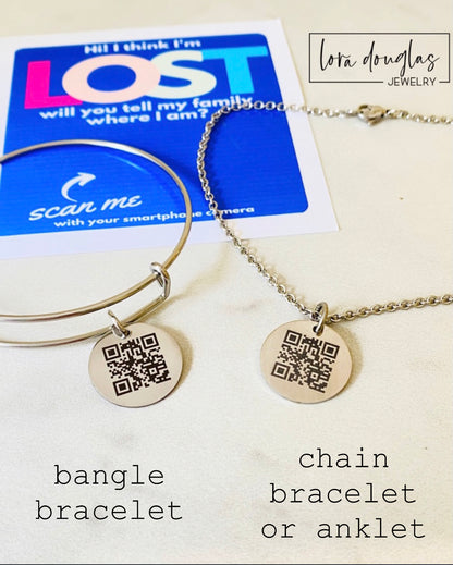 A QR code charm placed on a display stand, with a silver chain necklace that says 'Lost - will you tell my family I'm ok?' engraved on a blue disc.