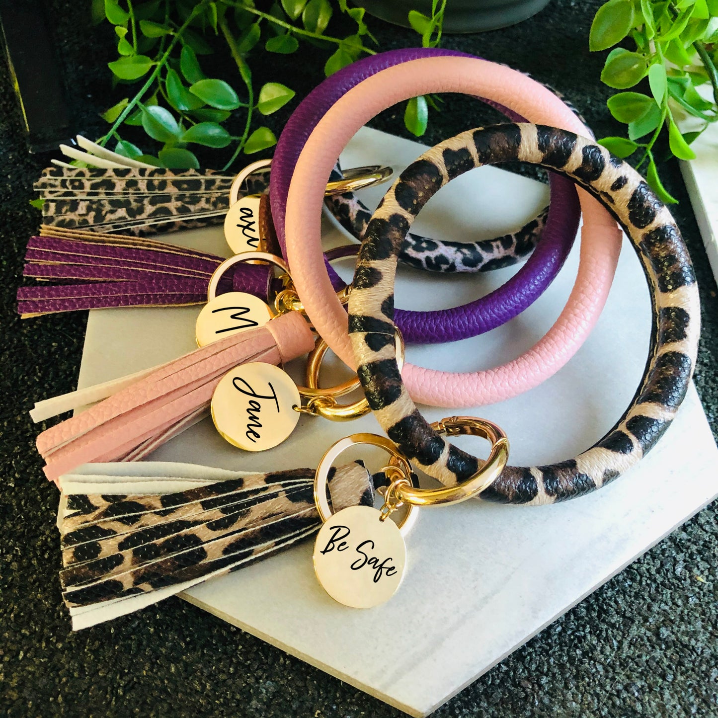 A leather keychain bangle bracelet with a leopard print design, featuring a gold stainless steel disc with engraved initials 'M&M', and a zipper closure.