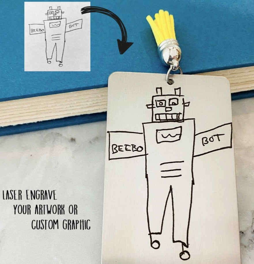 A custom stainless steel bookmark with a laser engraved child's drawing, presented next to its navy blue velvet drawstring pouch.