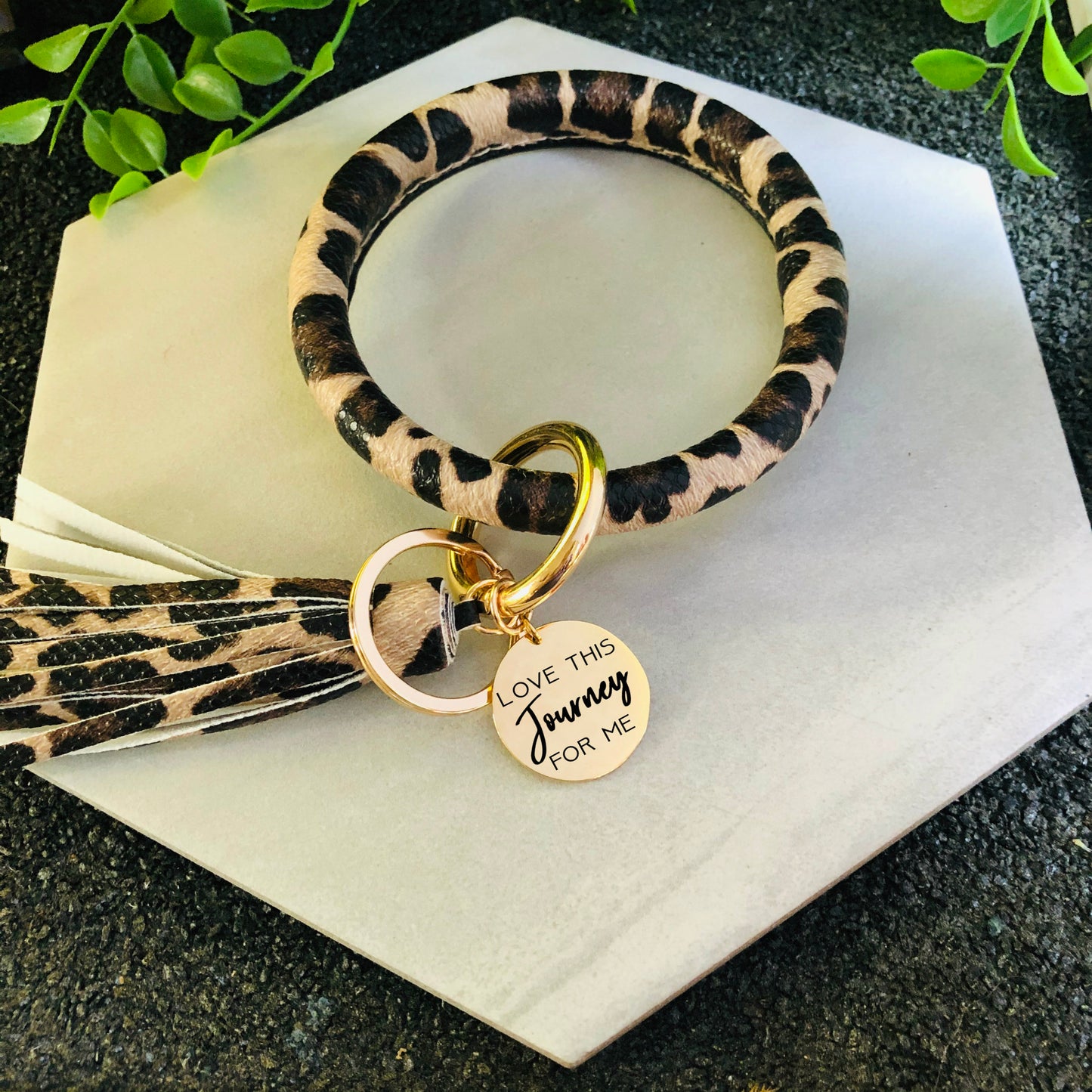A leather keychain bracelet with a gold stainless steel disc and the engraving 'Love This Journey For Me'.
