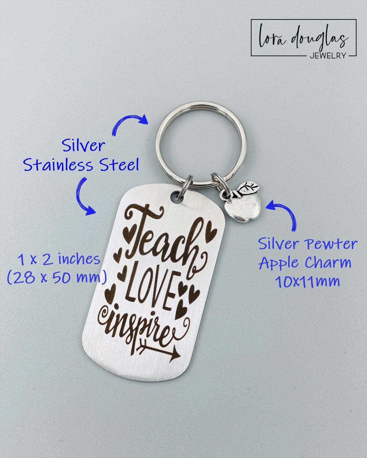 A silver stainless steel dog tag keychain with a silver pewter apple charm, engraved with the text 'Teach Love Inspire' accompanied by a heart symbol.