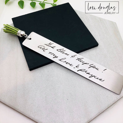 Two stainless steel bookmarks with engraved handwriting, with tassels, displayed on a wooden surface.