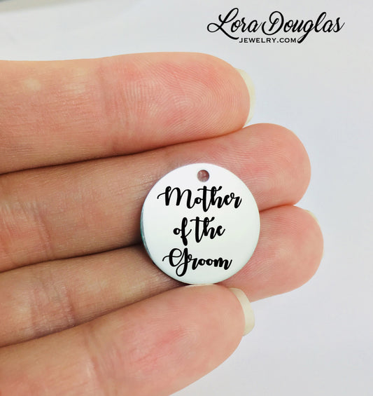 A round stainless steel pendant with the engraved text 'Mother of the Groom' held between fingers.