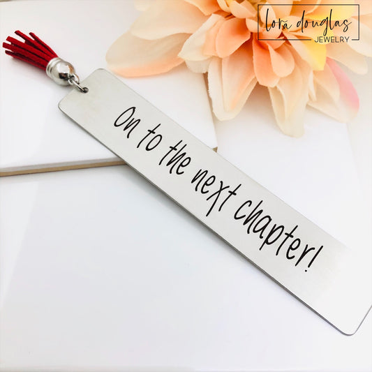 on to the next chapter bookmark engraved on stainless steel metal bookmark with leather tassel, inspirational graduation gift displayed inside open book with permanent laser engraving and brushed silver finish