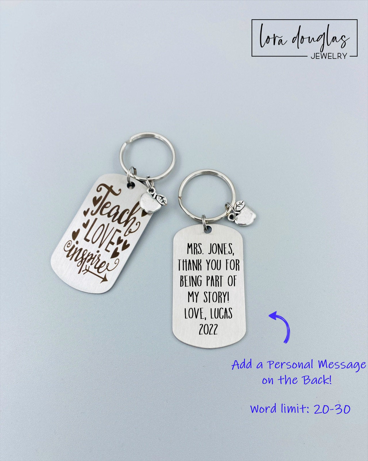 A silver stainless steel dog tag keychain with a silver pewter apple charm, engraved with the text 'Teach Love Inspire' accompanied by a heart symbol.