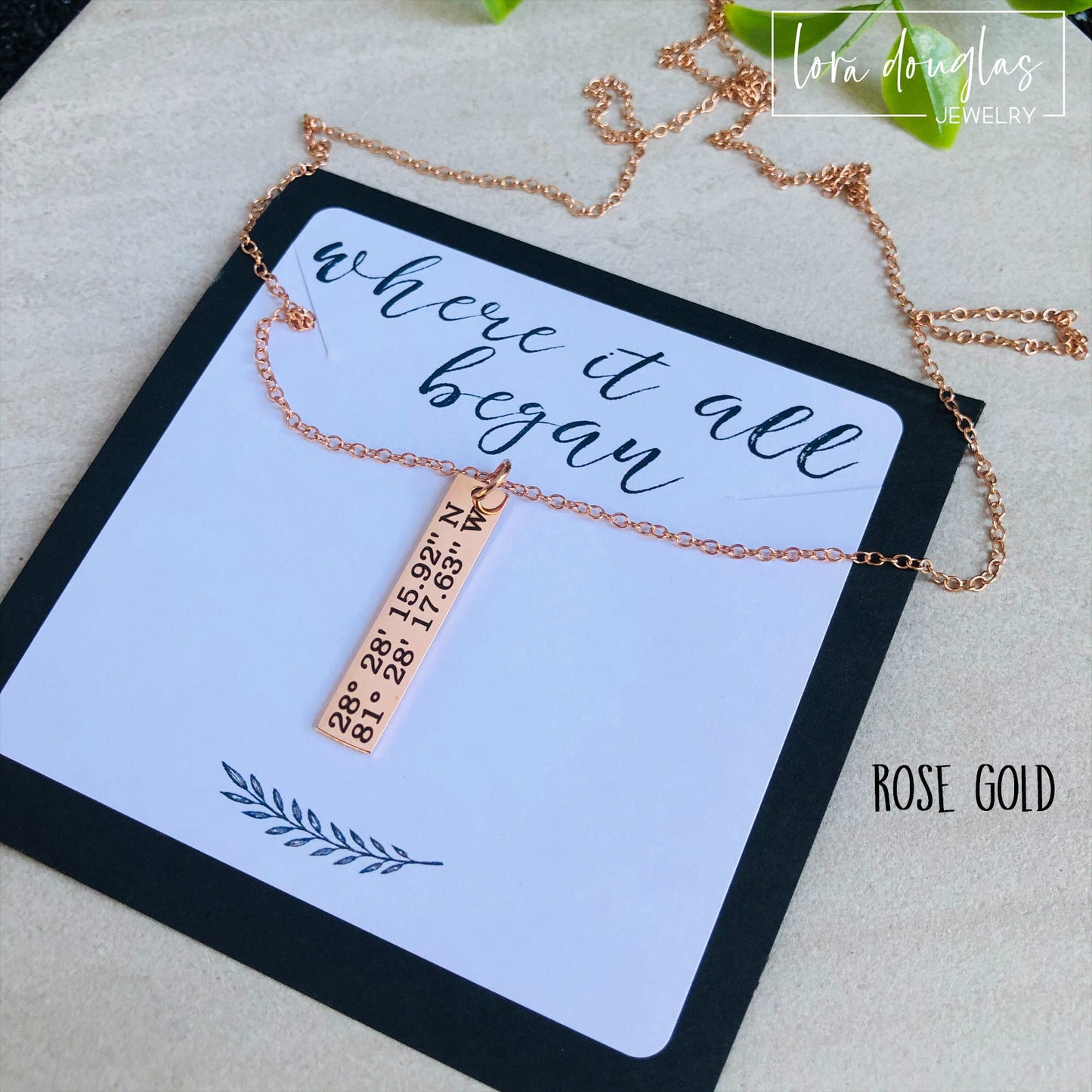 A rose gold necklace with a personalized engraving of a latitude and longitude on a pendant, displayed in a box with a blue and white design on the interior lining.