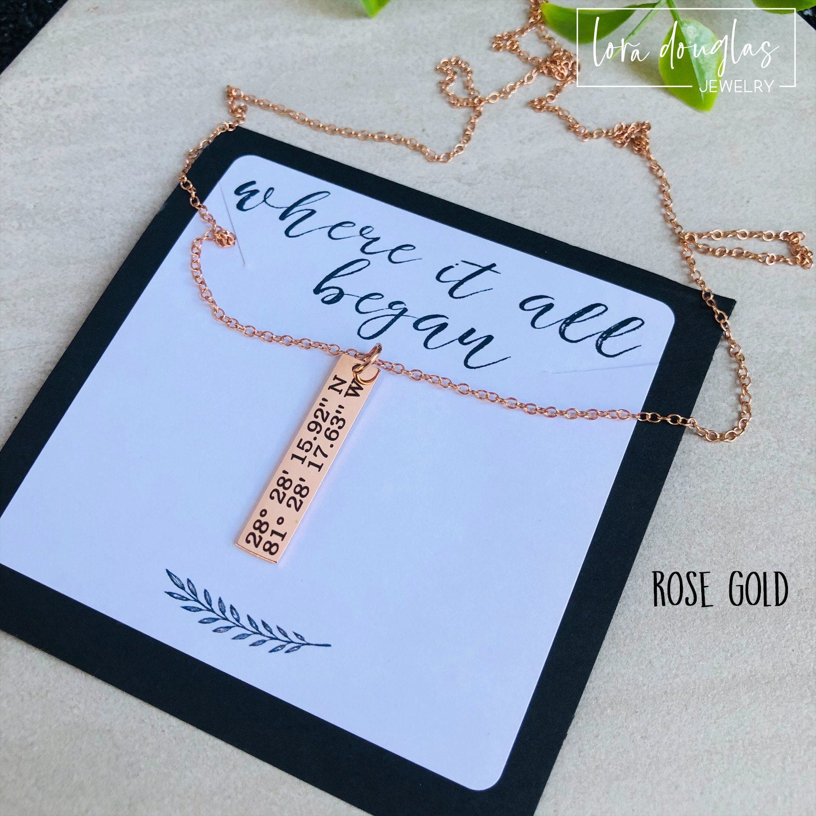 A rose gold necklace with a personalized engraving of a latitude and longitude on a pendant, displayed in a box with a blue and white design on the interior lining.
