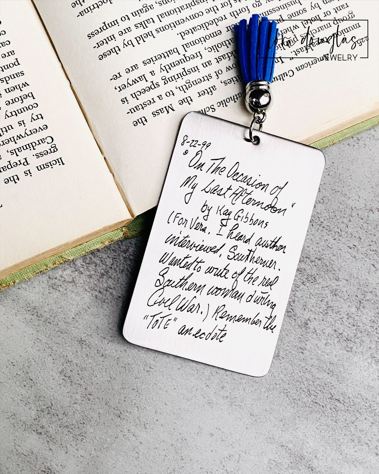 A custom engraved bookmark with a handwritten message engraved on the metal. The bookmark is silver with black engraving and has a navy blue Tassel.